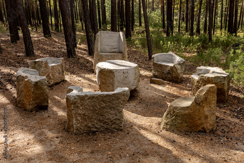 Rustic Stone Seating Area in a Pine ForestRustic stone chairs arranged in a circle in a peaceful pine forest, surrounded by tall trees, natural vegetation and warm sunlight. Atmospheric outdoor settin