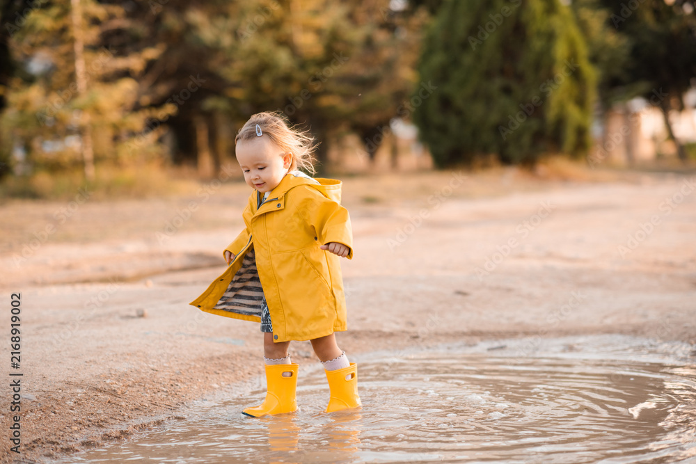 Fototapeta premium Cute kid girl 2-3 year old wearing yellow raincoat and rubber boots walking in puddles outdoors