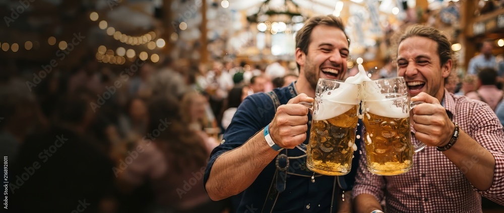Obraz premium Two men cheering with large beer mugs at a lively festival. Friends celebrating together in a traditional beer hall with festive lights and copy space.