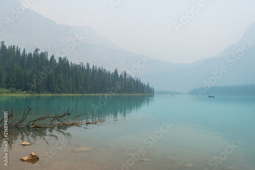 Emerald Lake, Canada - August 14, 2026: Emerald lake surrounded by forest and mountains under hazy sky caused by wildfire smoke. Canoes on the calm water create an atmospheric and moody summer scene.