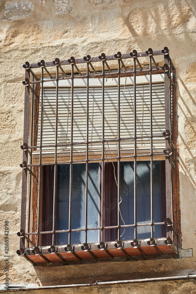 Obraz premium Traditional window with wrought iron security grille and wooden shutters on a weathered stone facade in Baeza, Andalusia, Spain