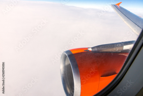 A view from an airplane window looking down past a metallic and red engine nacelle at a dense layer of fluffy white clouds. Vantage point, a commercial flight. An aerial photograph from high altitude.