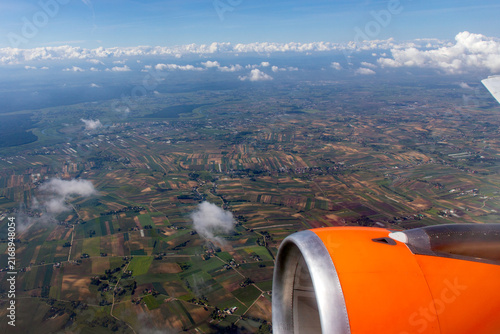 On the airplane an aircraft engine is visible from a window seat, looking down at an aerial view of green fields and patchwork farmlands. Patchwork agricultural landscape and scattered clouds below. 