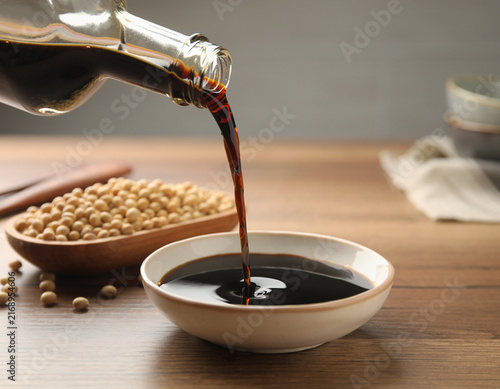 Dark soy sauce being poured from a glass bottle into a ceramic bowl, with dry soybeans in a wooden dish nearby. This kitchen setup for Asian cooking showcases a savory condiment ingredient for meals.