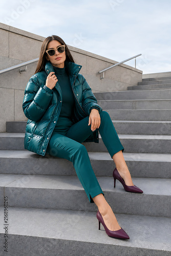 Outdoor Fashion Portrait of Stylish Woman in Teal Puffer Jacket and Sunglasses Sitting on Stone Steps