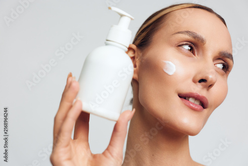 Obraz na plátně Bright woman applying moisturizer, close up of face with a pump bottle, soft stu