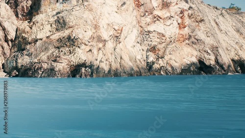 A rocky cliff stands tall beside a large body of water. The sun shines on the surface of the water, creating ripples. Trees are scattered on the cliff in the background.