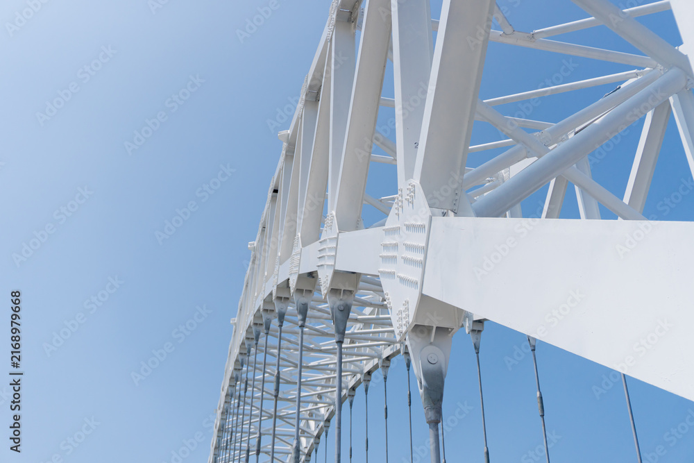 Obraz premium White steel bridge curves gracefully against clear blue sky. Strong structural beams form elegant, geometric pattern. Modern engineering meets natural sky in striking contrast. Ideal for architecture