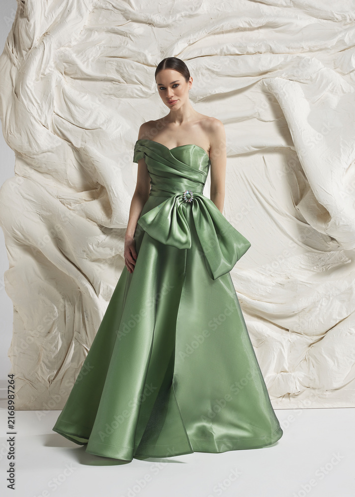 Fototapeta premium Full length shot of elegant woman wearing luxury sage green strapless satin ballgown with oversized bow at waist and wide hem posing gracefully against sculptural white studio wall backdrop
