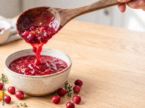 Cranberry Sauce Spoonful: A wooden spoon lifts a dollop of glistening cranberry sauce, flecked with orange zest, from a rustic bowl, garnished with fresh cranberries and herbs.