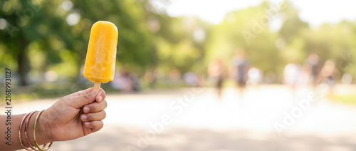 Child holding yellow popsicle in summer park with blurred background of people walking