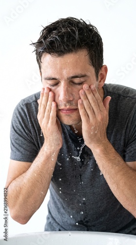 A man washing his face with water in front of a white background. He has his eyes closed and his hands are rubbing his face