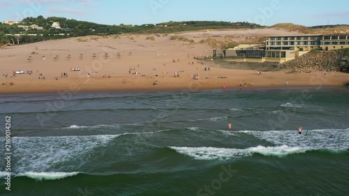 Visitors enjoy beach activities at Guincho Beach in Portugal aerial view. Surfers ride the waves while others relax on the sand during the afternoon sunlight.