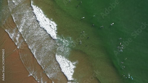 Surfers catch waves at a California beach aerial view. Ocean is clear, and the sun shines bright. People enjoy the beach while others paddle out to surf.