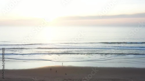 People walk along the beach as the sun sets over the ocean. Waves gently roll onto the shore. The sky shows colors as the day comes to an end.