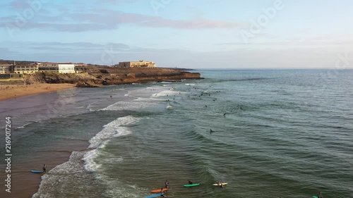 Waves break at Carcavelos Beach as surfers ride them under a clear sky at sunset aerial view. People enjoy surfing and swimming as the day winds down.