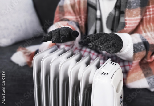 Fotografie Freezing at home in winter, a person wrapped in a blanket is warming their cold hands in gloves over an electric radiator heater