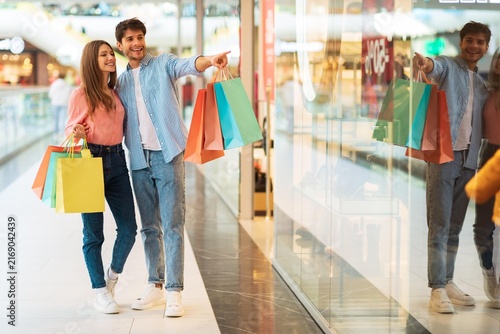 Canvastavla Smiling Young Couple With Shopping Bags, Embracing And Pointing At Store Window In A Shopping Mall, Full Of Joy And Excitement While Enjoying Their Day Out Shopping Together