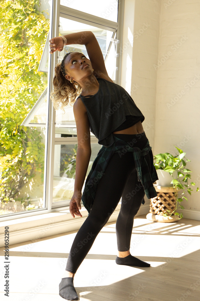 Naklejka premium Adult African American woman stretching near open window on sunlit floor, plaid shirt tied, planter