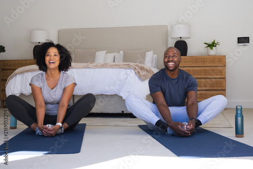 African American couple sitting and stretching on navy yoga mats in bedroom with water bottles