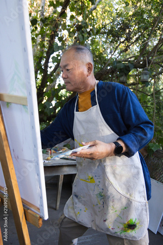 Obraz na plátne Senior Asian man painting at garden patio, holding palette and brush by easel, w