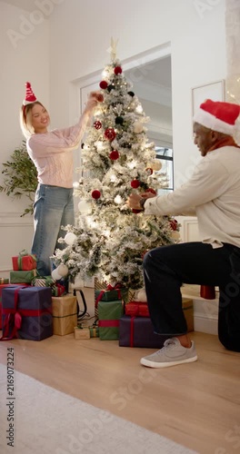 Vertical video: Couple wearing hats decorating tree at home after holiday cue, placing red baubles