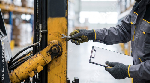 Plakat Warehouse worker in uniform holding a wrench and a clipboard while performing maintenance on a forklift in a distribution center