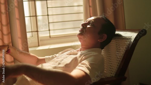 Senior Indian man resting comfortably in an armchair beside a bright window at home.