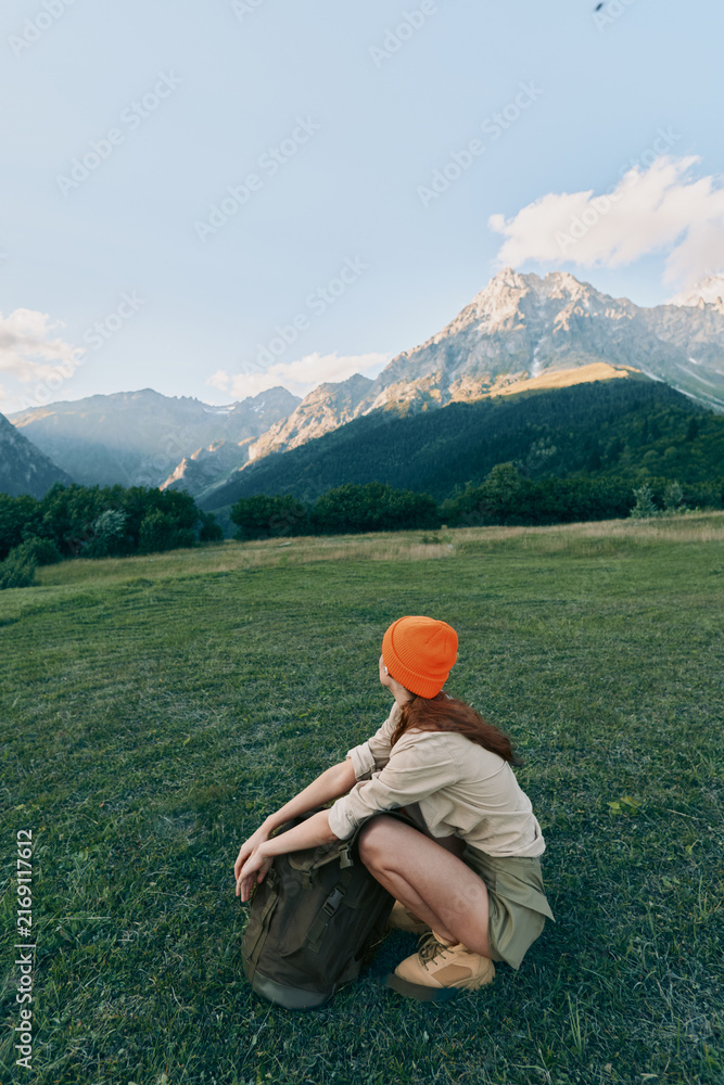 Fototapeta premium Woman sits in a wide grassy field with a backpack and orange beanie, enjoying a peaceful outdoor lifestyle amid distant mountains and expansive nature scenery.