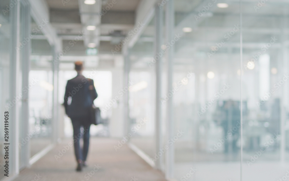 Obraz premium Professional worker walking inside a modern office surrounded by glass partitions and bright work areas. The blurred environment provides a clean background for advertising and business content.