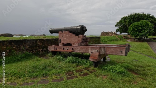 Wide shot of an old cannon on a wooden carriage near a historic fort in rainy, overcast Galle with a stormy ocean visible in the distance.