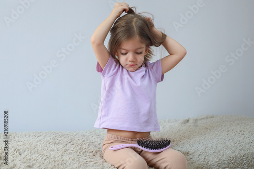 Young girl sitting on soft rug combing hair with pink brush, wearing lilac shirt and beige pants, calm indoor grooming moment against plain light background, with girl, child, hair