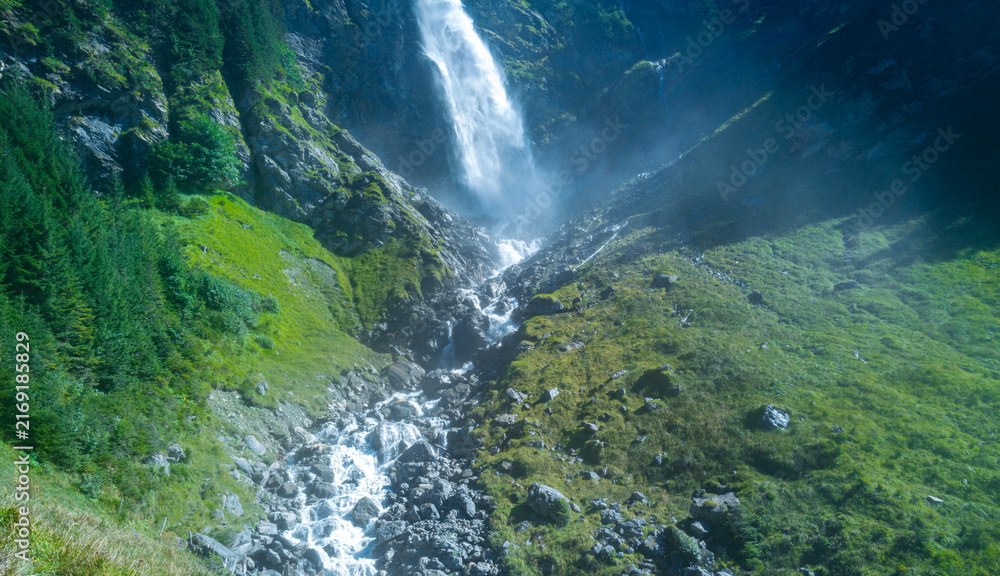 Fototapeta premium Waterfall cuts through a rocky gorge. Waterfall rushes down dark alpine rocks. Mountain landscape contains waterfall inside wilderness. Swiss nature surrounds waterfall near gorge.