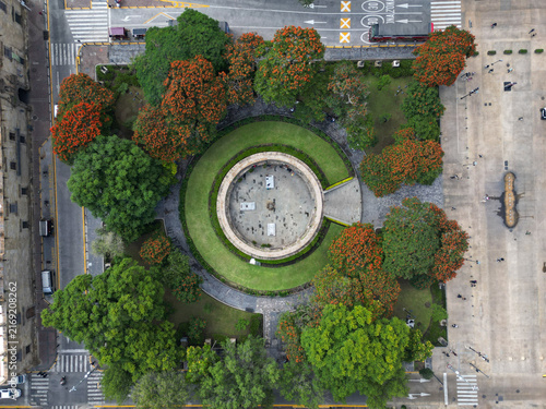 Aerial top-down shot of the Rotunda of the Illustrious Jaliscans in Guadalajara, Mexico. Green park with orange flowering trees, urban roads.