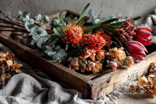 Elegant floral arrangement featuring vibrant protea flowers, eucalyptus, and dried elements on a rustic wooden tray with soft, neutral background.