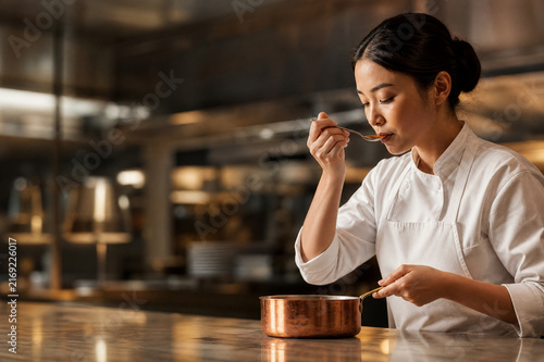 Female chef tasting sauce from a copper pot with a spoon in a warmly lit restaurant kitchen