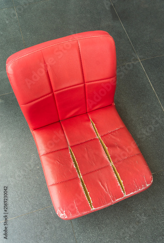 High-angle view of an old, broken red floor chair with severely torn faux leather. The ripped seams reveal the inner foam padding, highlighting neglect and a desperate need for repair.
