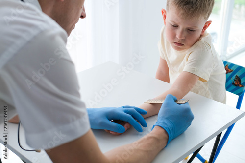 Close-up photograph of a child's hand with a red spot reaction to a Mantoux test 72 hours after injection. A nurse in blue gloves uses a ruler to check the reaction.
