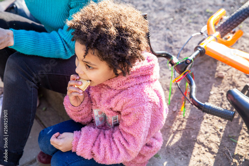 Young multirracial daughter eating snack next to mother and bicycle