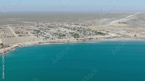 Aerial view of coastal town with turquoise ocean and desert landscape beyond.