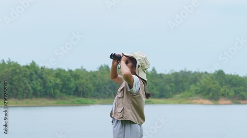 Asian kid wearing safari hat looking through binoculars by the lakeside