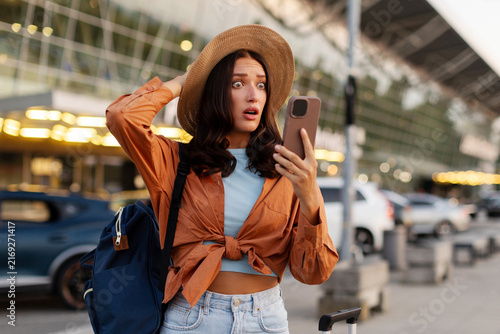 Shocked young woman looking at smartphone while standing with backpack and suitcase outside modern airport terminal