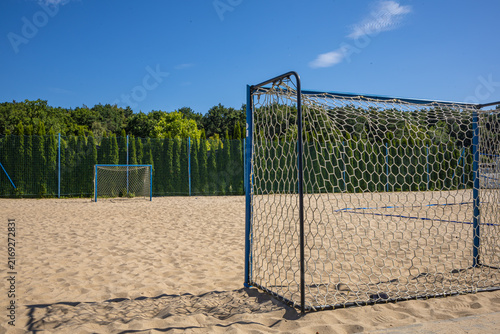 Beach Soccer Goal on Sandy Recreational Sports Field. A full-size beach soccer goal with a net stands on a sandy outdoor sports field surrounded by green woodland. The empty goal and open sandy playin