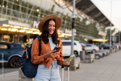 Картината върху платно Smiling lady using her smartphone while holding passport, standing with suitcase