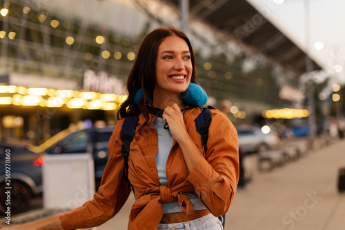 Joyful woman tourist wearing comfortable neck pillow and carrying backpack while preparing to board flight at airport