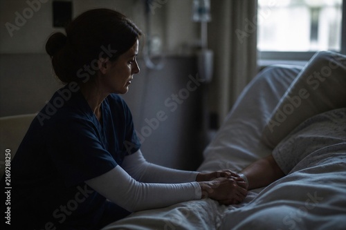 Woman nurse holding elderly patient hand in bedroom. Compassionate home healthcare, hospice support and caregiving concept.