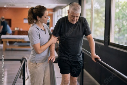 Woman physiotherapist assisting senior man with walking exercise in rehabilitation clinic. Recovery training, mobility support, physical therapy and healthy aging concept.