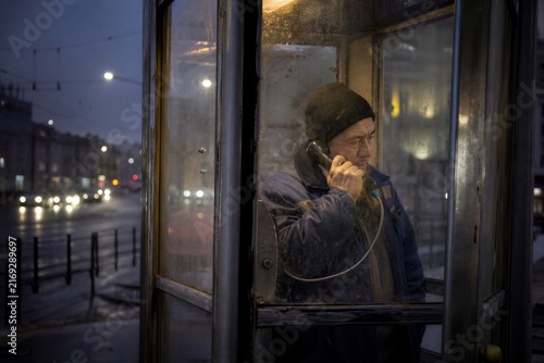 Man making a phone call inside a weathered street telephone booth at dusk. Urban communication, solitude and migrant worker journey concept with city traffic lights.