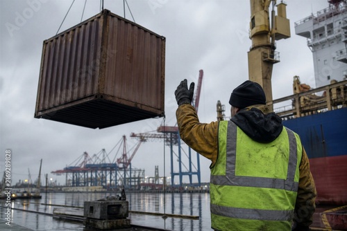 Dockworker guiding a suspended cargo container at a busy seaport. Maritime logistics, industrial safety and global shipping operations on an overcast day.