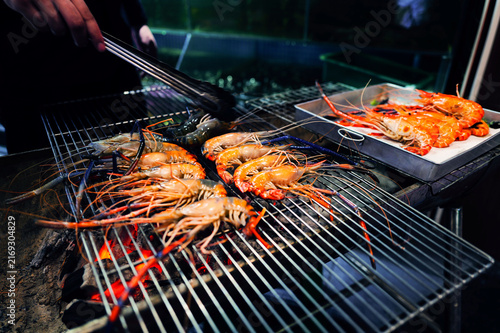 Foto Chef hand using tongs to grill giant river prawns over flaming charcoal stove at Thai night street food market